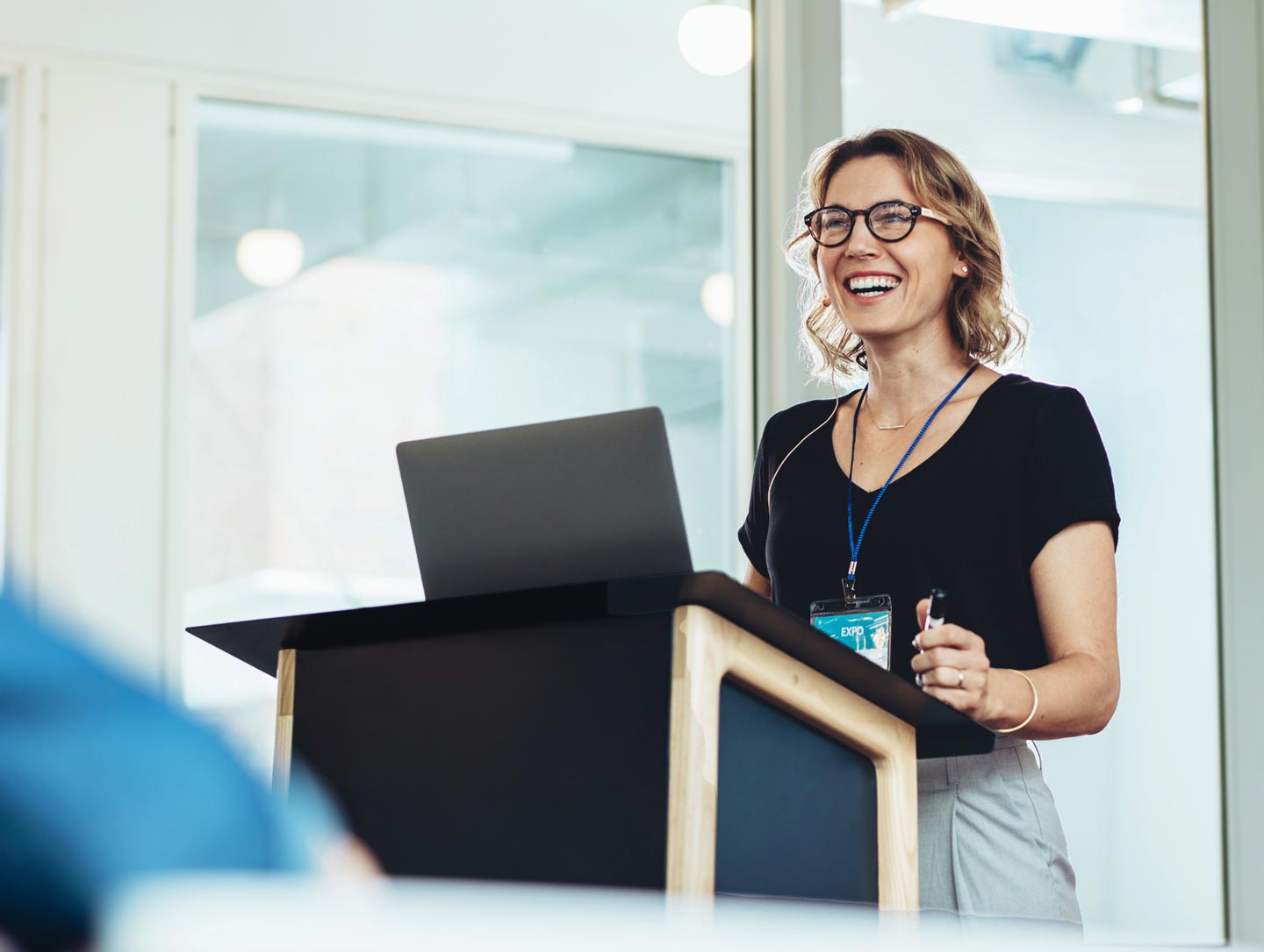 A smiling female industry speaker with glasses addresses an audience from a wooden podium during a business convention.
