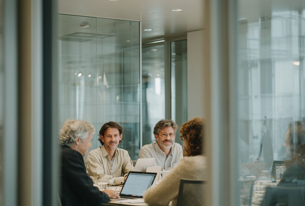 A professional business team collaborating around a laptop computer in a modern corporate glass conference room.